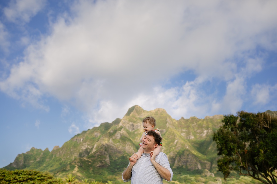 dad carries baby daughter on his shoulders with kualoa mountains in the background during a family photo session in oahu